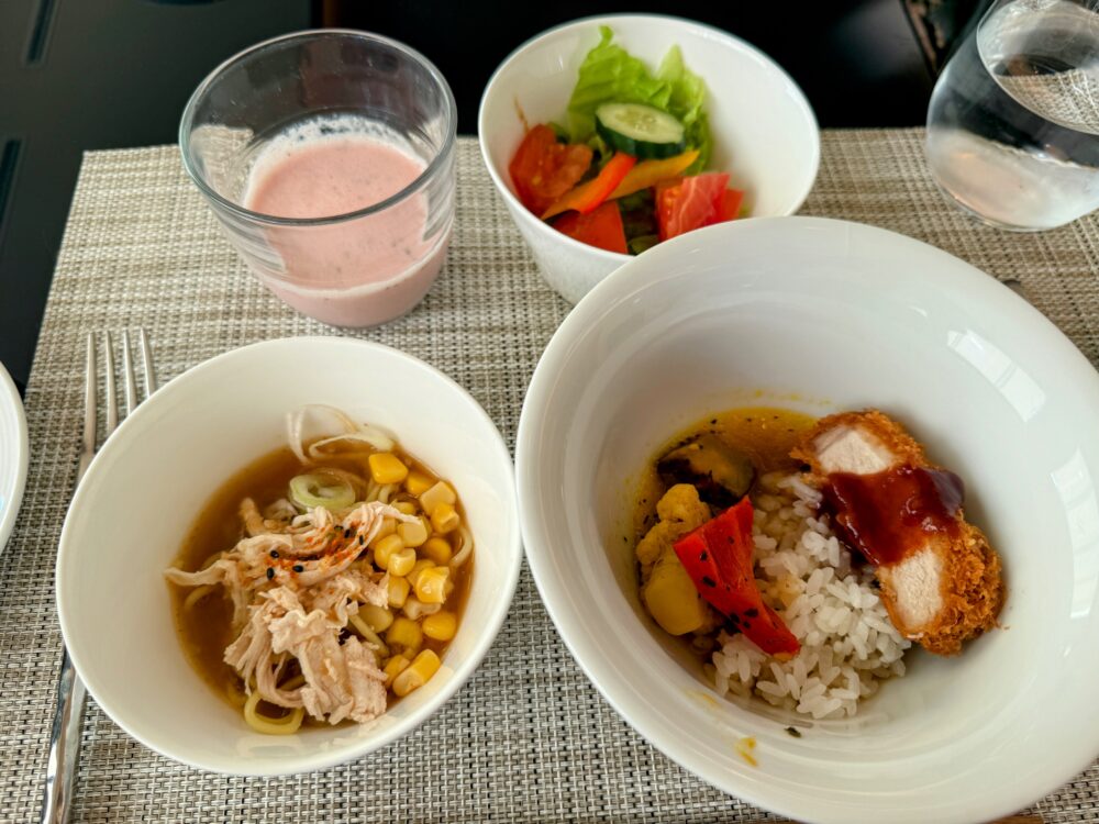 ホテルの朝食メニュー。白いボウルに盛られたチキンラーメン、カツカレー、サラダ、そしてピンク色のスムージーが、麻目調のランチョンマットの上に置かれている。