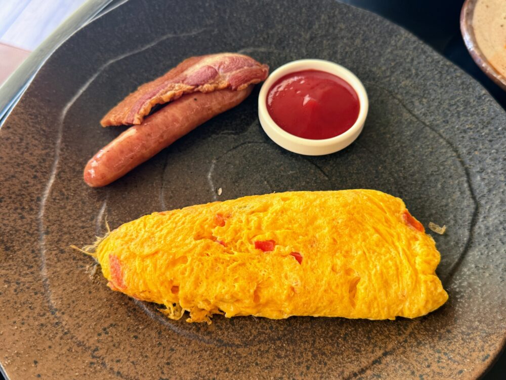 朝食のメインディッシュ。ダークブラウンの陶器のプレートに、プレーンオムレツ、ソーセージ、ベーコン、そして小鉢に入ったケチャップが盛り付けられている。シンプルで洗練された洋食メニュー。