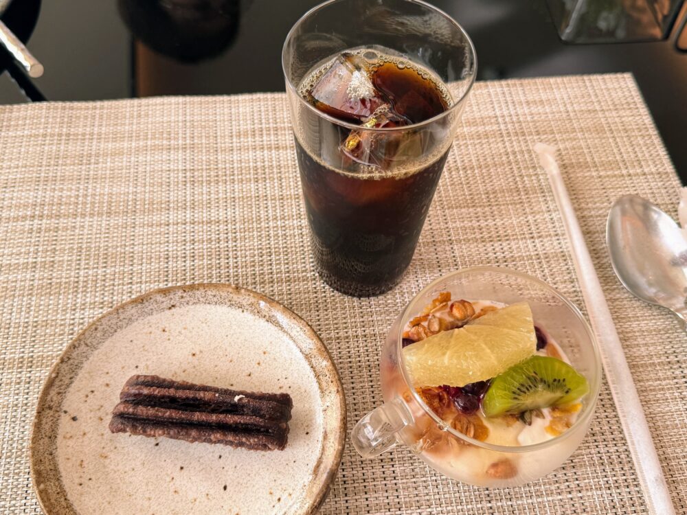 ホテルの朝食のデザートとドリンク。陶器のプレートに乗ったチョコレートチュロス、グラスに入ったフルーツヨーグルト(グラノーラ、グレープフルーツ、キウイ)、そしてアイスコーヒーが並んでいる。優雅な朝のカフェタイムを思わせる。