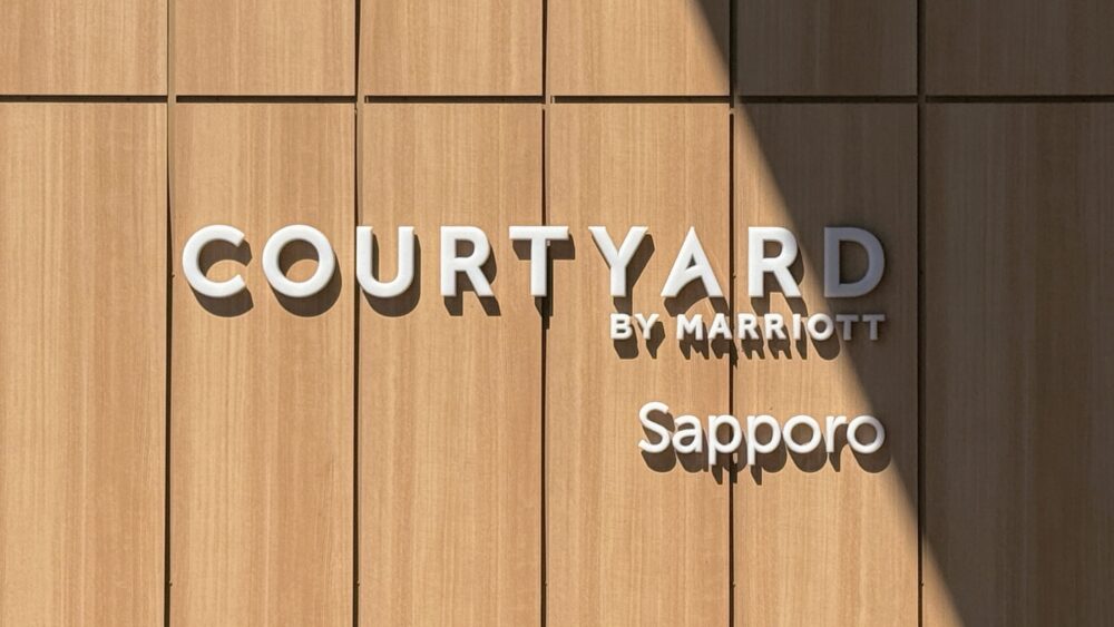 ホテルの木製壁面に設置された「COURTYARD BY MARRIOTT Sapporo」の白い立体的なホテルロゴ看板。日差しと影が当たっている。
