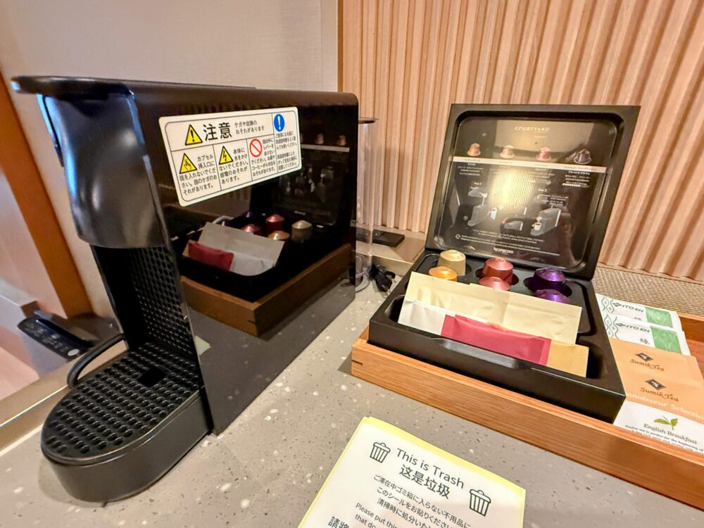 客室に設置された黒いカプセル式コーヒーメーカーと、ネスプレッソのカプセル、お茶のティーバッグが収納されたボックスのクローズアップ。注意事項のステッカーも貼られている。
