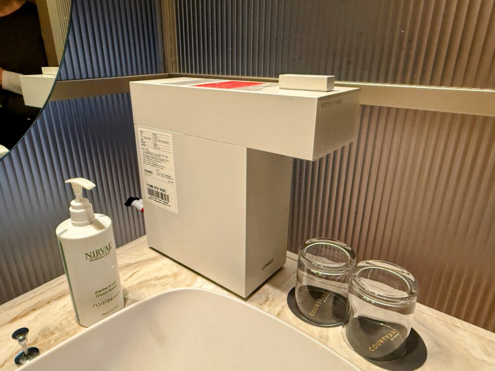 客室の洗面台に設置されたCOWAY製の浄水器（ウォーターサーバー）と、隣にあるNIRVAEのハンドウォッシュボトル、COURTYARDのロゴ入りグラス。