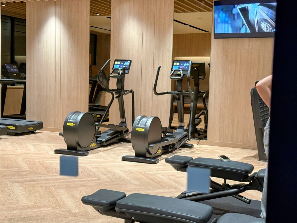 ホテルのフィットネスセンターの様子。Technogym製のクロストレーナー（エリプティカル）やランニングマシンが木目調の壁を背景に配置されている。