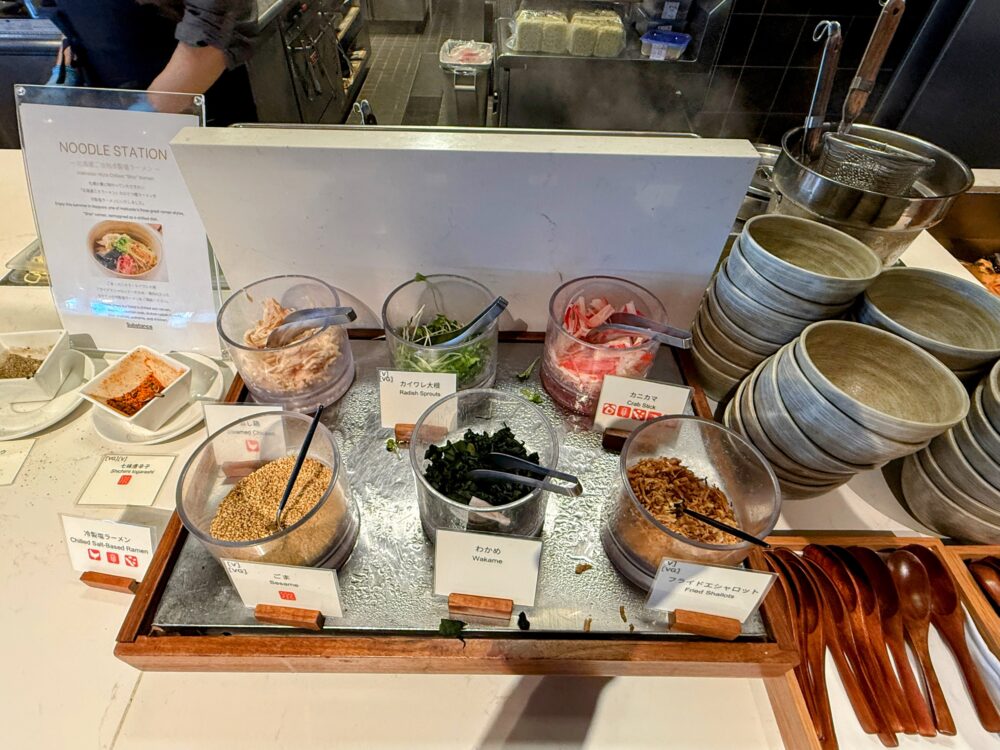 レストラン「Substance」のヌードルステーション（北海道ご当地冷製塩ラーメンのトッピングコーナー）。鶏むね肉、カイワレ大根、カニカマ、ごま、わかめ、フライドエシャロットなどの具材が並ぶ。