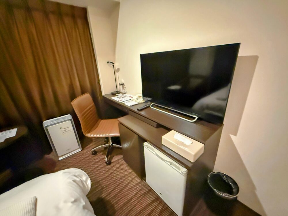 客室のデスクと大型テレビ、冷蔵庫。奥にカーテンと空気清浄機