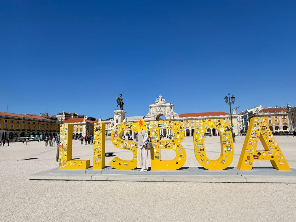 コメルシオ広場を背景に、「LISBOA」と書かれた黄色の大きなモニュメントと記念撮影する旅行者