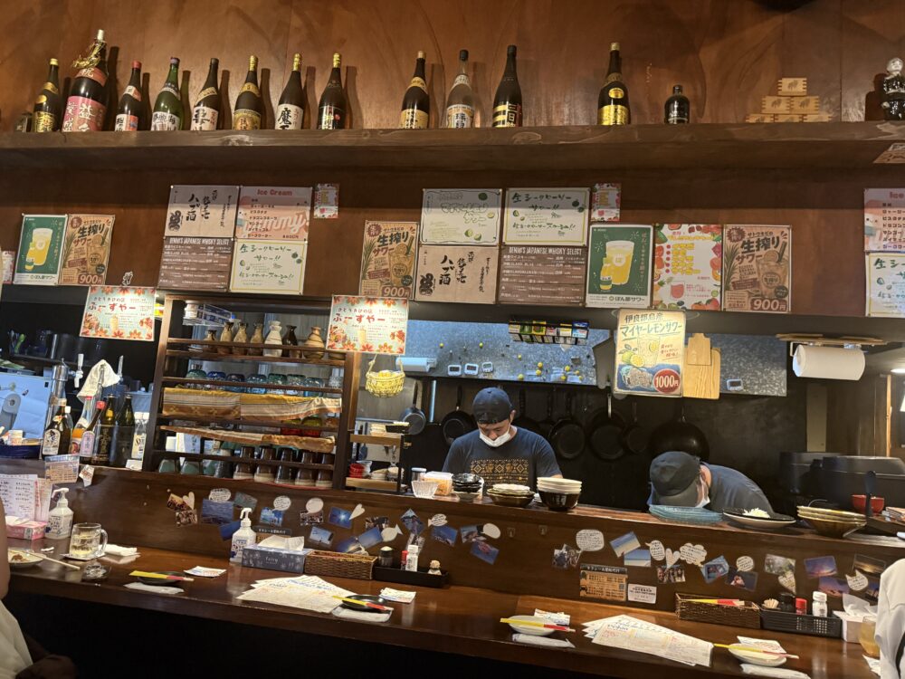 居酒屋でんみのカウンター。棚に泡盛などのボトルが並ぶ。
