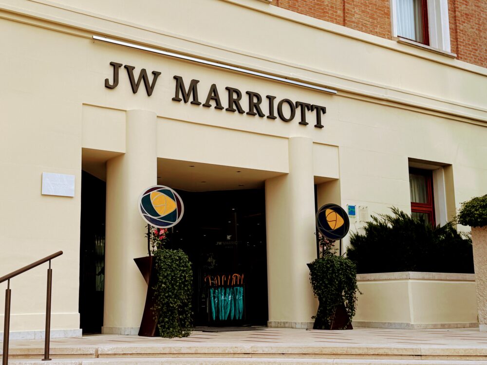 「JW MARRIOTT」の立体文字が掲げられたJWマリオット・ベニスの陸側エントランス。マリオットボンヴォイのロゴを描いたステンドグラス風の丸いオブジェと蔦の植栽が入口の左右に置かれ、奥にはターコイズブルーの傘立てが見える