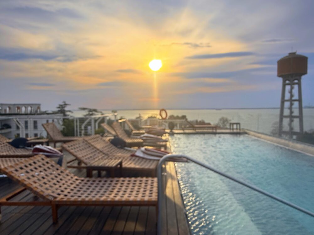 夕暮れ時の屋上インフィニティプール。ラグーンの水平線に沈む美しい夕日が水面に反射し、手前のデッキチェアを黄金色に染めている