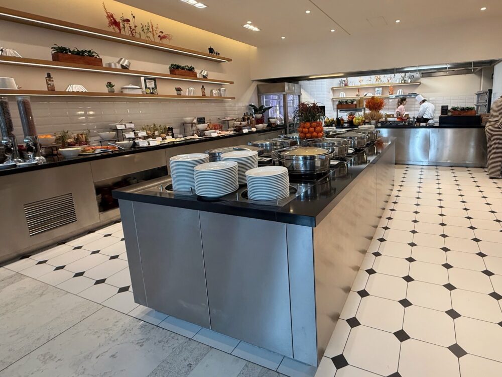 朝食会場「Cucina」のビュッフェコーナー全景。中央のアイランドテーブルに温かい料理、壁沿いにはパンやシリアルなどが美しく並ぶ広々とした空間