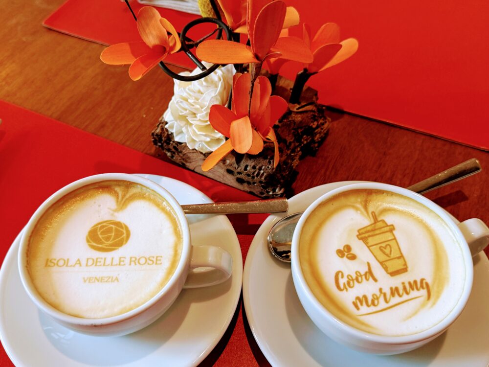 「ISOLA DELLE ROSE VENEZIA」のロゴと「Good morning」の文字、コーヒーカップのイラストが描かれた、見事なラテアートの2杯のカプチーノ