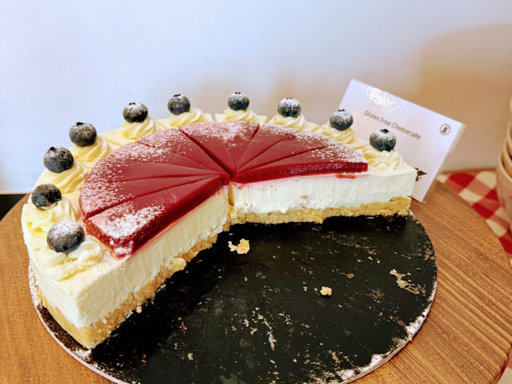 ホール状のグルテンフリー・チーズケーキ（Gluten Free Cheesecake）。表面の赤いゼリー層と、ブルーベリーとクリームのトッピングが美しい
