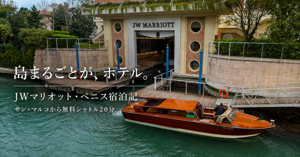JWマリオット・ベニスの水上エントランス。「JW MARRIOTT」の文字が掲げられたレンガ造りの建物に木製の水上タクシーが横づけされ、手前にはエメラルドグリーンのラグーンと木製の桟橋が広がる。「島まるごとが、ホテル。JWマリオット・ベニス宿泊記／サン・マルコから無料シャトル20分」の文字入りアイキャッチ画像